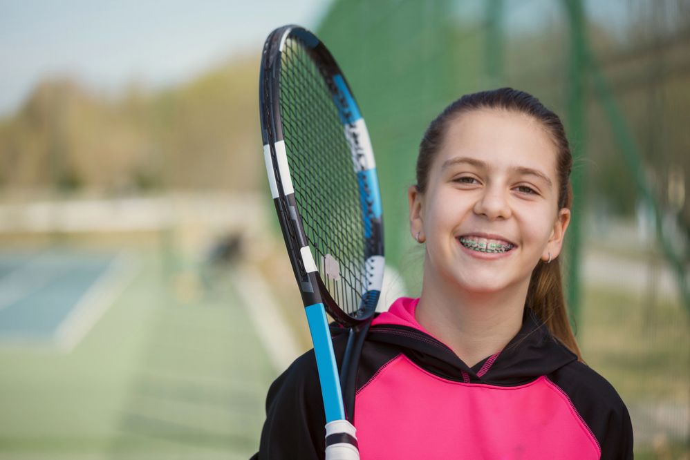 A smiling young girl with braces holding a tennis racket on an outdoor tennis court.
