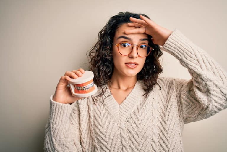 A woman in glasses and a beige sweater looks worried as she holds a dental model with braces.